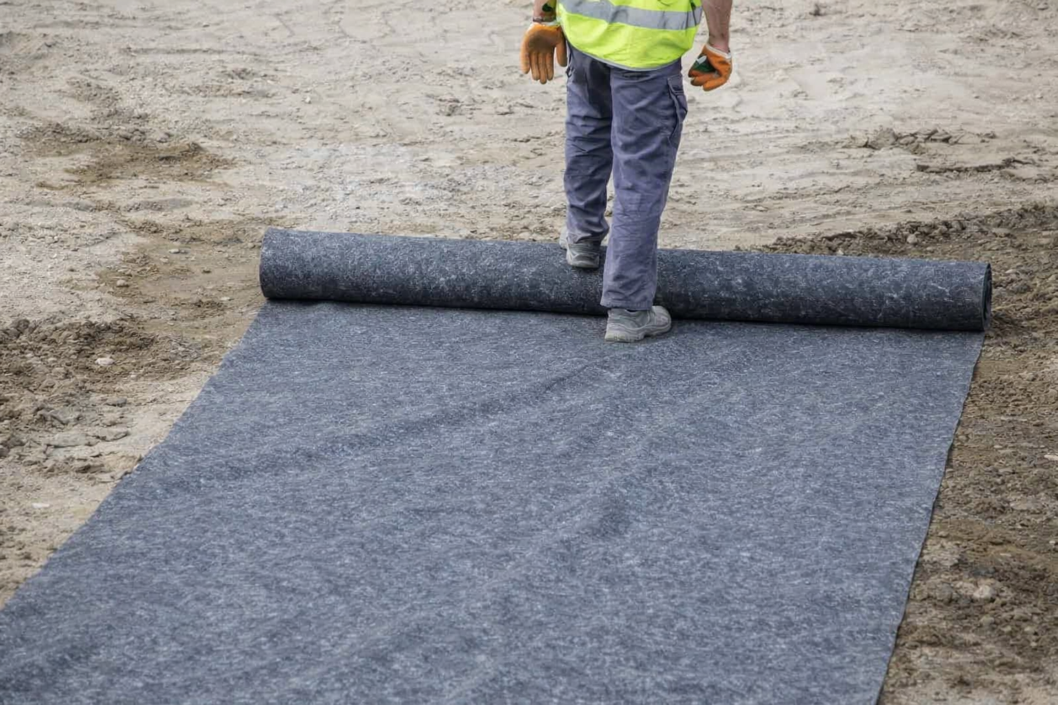 Pokládka netkané geotextilie na upravený podklad – pracovník v reflexní vestě rozvíjí roli geotextilie na stavbě